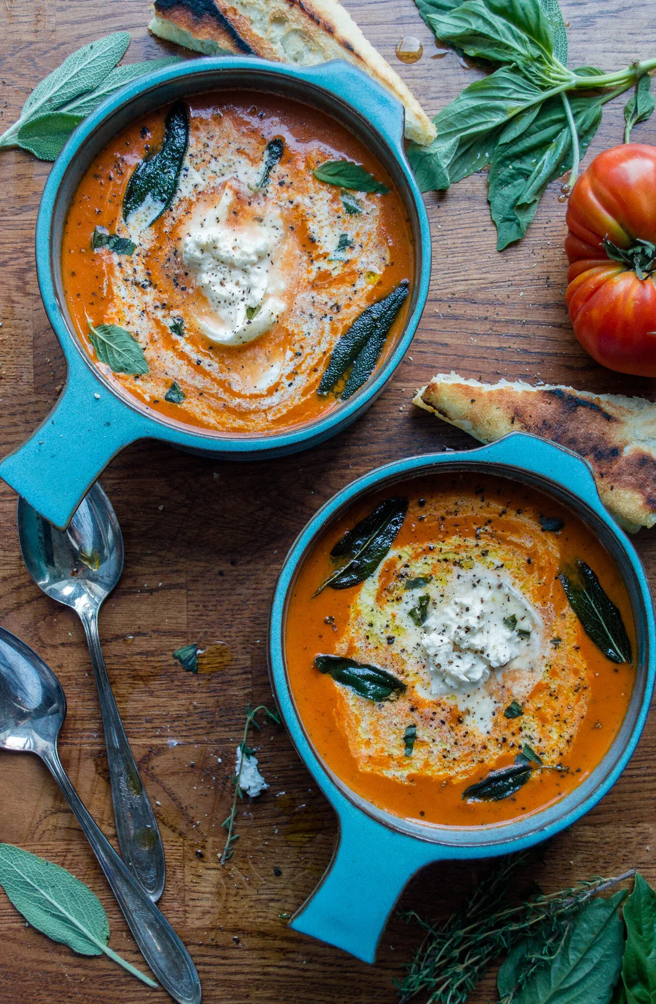 Overhead view of two bright teal crocks of Roasted Tomato and Red Pepper Bisque with Burrata and Garlic Tostada on a warm wooden board
