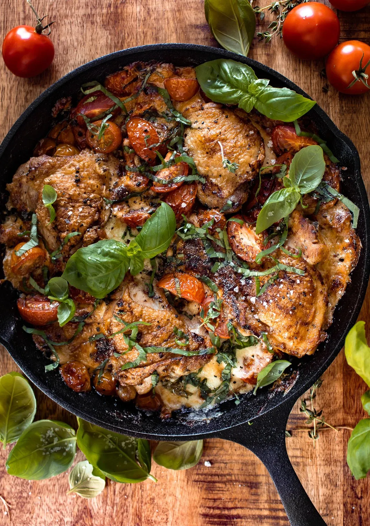 Overhead view of cast iron caprese chicken thighs in a black cast iron skillet on a rustic wooden board