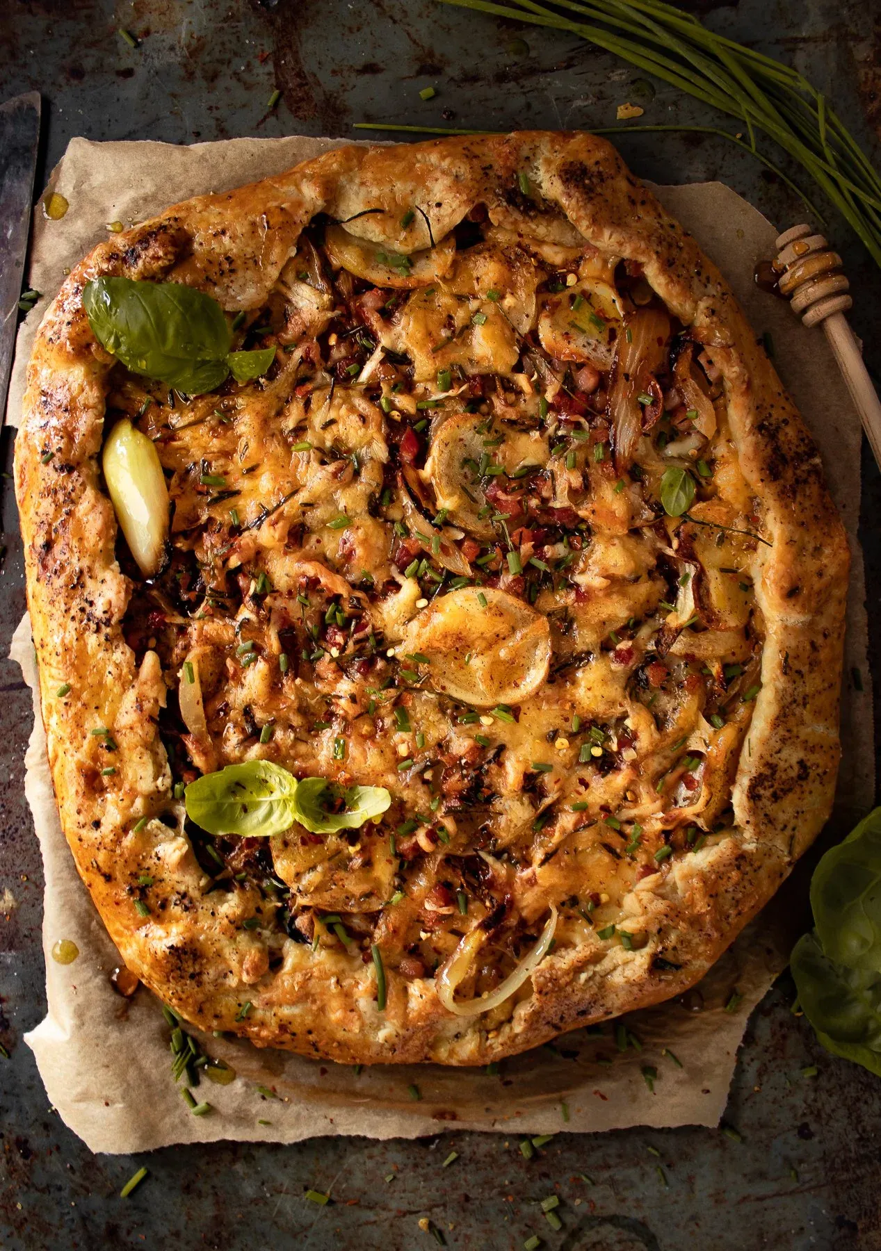 Overhead view of a whole cheesy potato, onion and chive tart with pancetta on parchment paper on a dark metal baking sheet