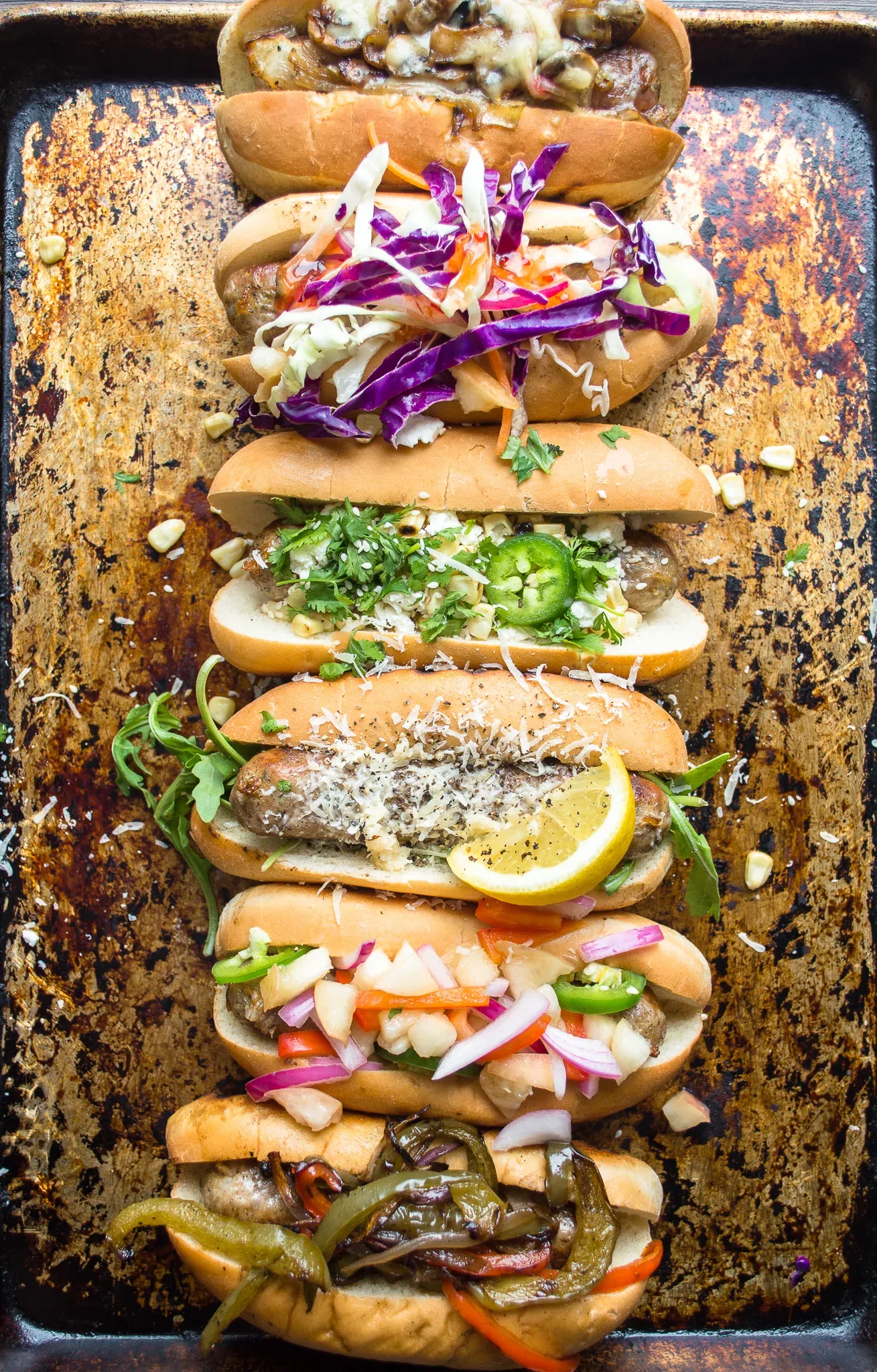 Five Better Than Basic Sausage Dogs lined up on a dark seasoned sheet pan, each with a different gourmet topping