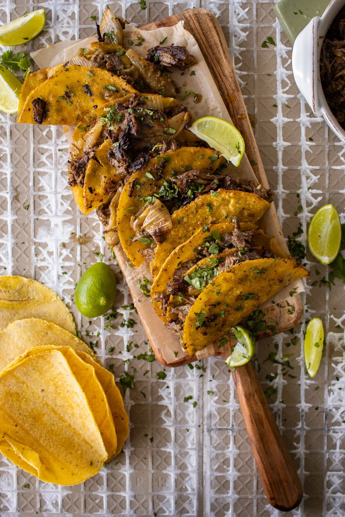 Eight Crispy Dominican Beef Tacos lined up on a long narrow wooden paddle board over parchment paper